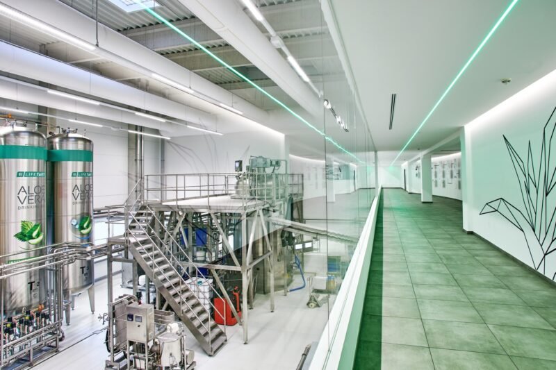 Couloir vitré au sein du site de production LR Health & Beauty, vue sur les cuves Aloe Vera et ambiance épurée.