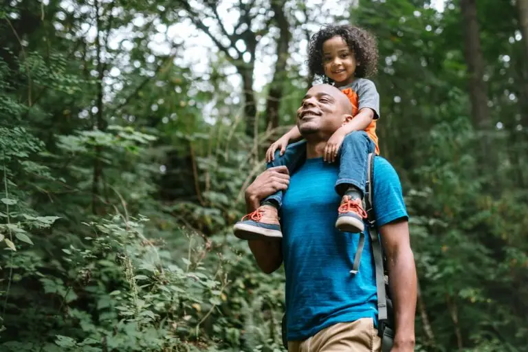 Un père portant sa fille sur ses épaules lors d’une promenade en forêt, incarnant la transmission, la joie et la connexion à la nature.