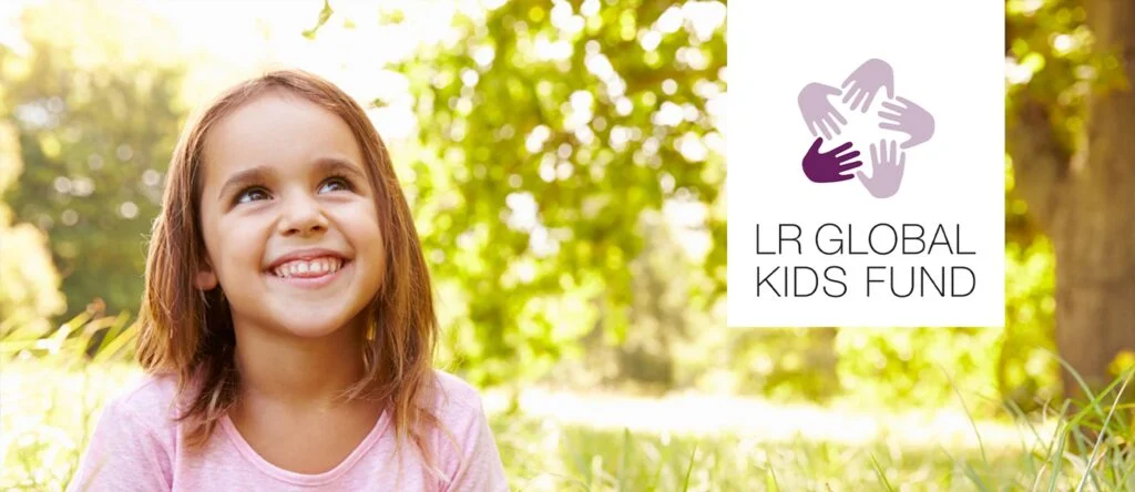 Niña sonriente sentada sobre la hierba, bajo un árbol – imagen que refleja esperanza y alegría.